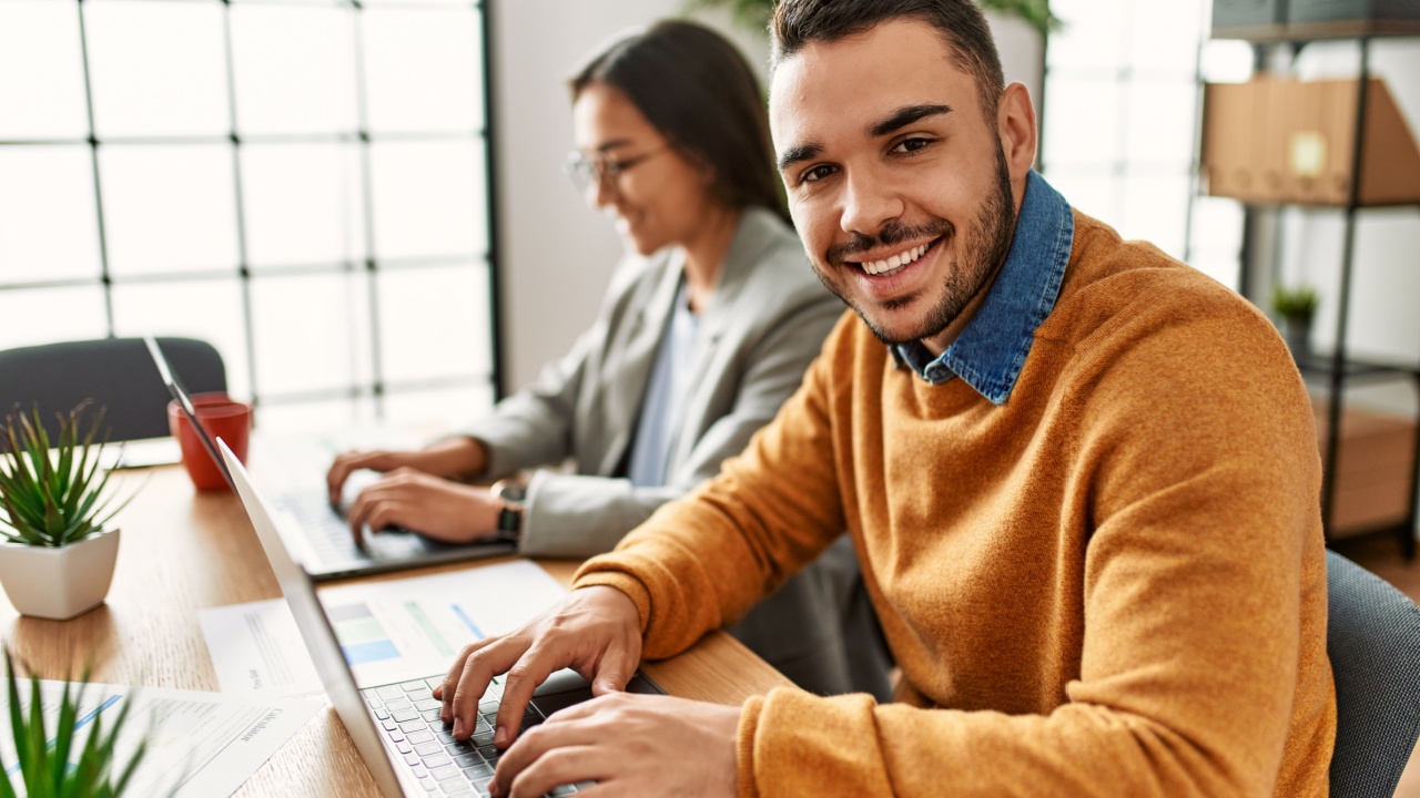 Smiling male employee working at computer with female co-worker in the background